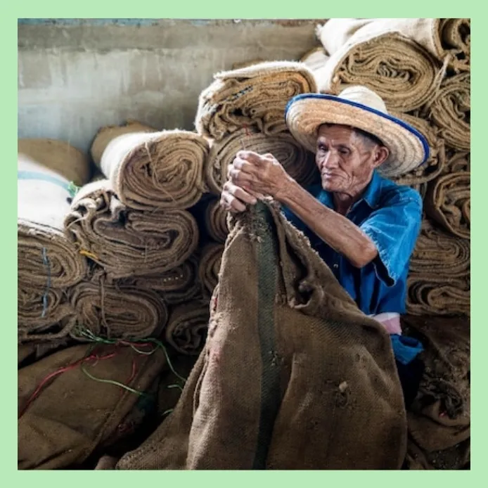 Man stitching used jute sacks beside rolls of burlap fabric